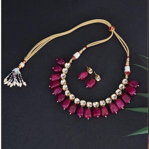 Könnte beinhalten: Ein Halsketten- und Ohrring-Set mit tiefroten, tropfenförmigen Edelsteinen. Die Halskette hat eine goldfarbene Kette und ist mit kleinen weißen Perlen verziert. Die Ohrringe spiegeln das Design der Halskette wider. Das Schmuckstück ist vor dunklem Hintergrund.