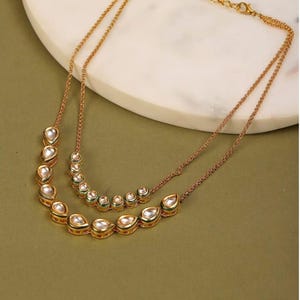 Puede incluir: Dos collares dorados con colgantes transparentes en forma de lágrima. Los colgantes tienen bordes dorados. Los collares están superpuestos, uno con colgantes más grandes y el otro más pequeños. El fondo es verde claro.