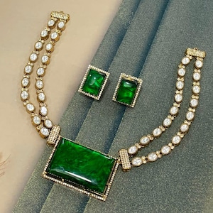 Puede incluir: Un conjunto de collar y pendientes a juego en tono dorado. El collar presenta un colgante rectangular grande de piedra verde y dos hilos de piedras blancas ovaladas. Los pendientes son piedras verdes cuadradas con marcos dorados. El conjunto de joyas se exhibe sobre una superficie gris.