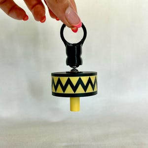 May include: A black and yellow bottle stopper with a geometric design. The stopper has a black ring handle and a yellow base. The design features a black zigzag pattern on a yellow band. The stopper is held up by a hand.