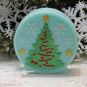 May include: A circular, pale blue soap featuring a Christmas tree design. The tree is green with red detailing, surrounded by white snowflakes and yellow stars. A festive holiday item.