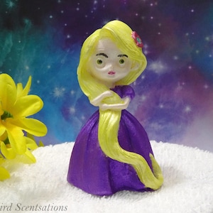 May include: A figurine of a character with long yellow hair, wearing a purple dress, and a pink flower in her hair. The figurine is standing on a white surface, with yellow flowers and a galaxy background.