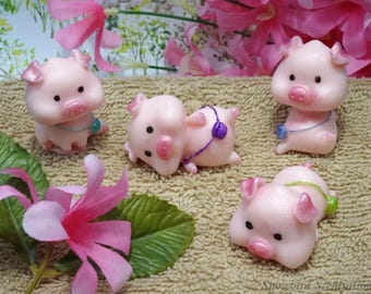 May include: A collection of five small, pink pig figurines with black eyes and pink snouts. Some pigs have blue or purple accents. The figurines are arranged on a textured, tan surface, with pink flowers and green leaves in the background.