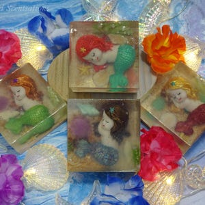 May include: Handmade mermaid soaps in clear square blocks, each featuring a sleeping mermaid with different colored hair and tails. The soaps are surrounded by seashells, flowers, and fairy lights, creating a whimsical, ocean-themed display. The text "Snowbird Scentsations" is visible.