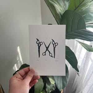 May include: A white card with a black scissor design is held in front of a large green plant. The scissor design includes three pairs of scissors, with the word "CUT" and other text below the scissors.