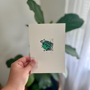 May include: A white card featuring a green and black turtle illustration on a green circle. The text "let them waddle" is printed below the turtle. The card is held in front of a plant in a woven basket.