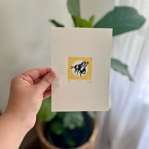 May include: A cream-colored print featuring a black and yellow bee design. The bee is set against a yellow square with a white circle. The print includes the words "Bee" and "Bloom".