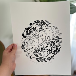 May include: Black and white linocut print featuring a running hare encircled by leaves. The artwork, titled "Hare and Hedge," is signed and numbered 13/50. The print is on a white background.