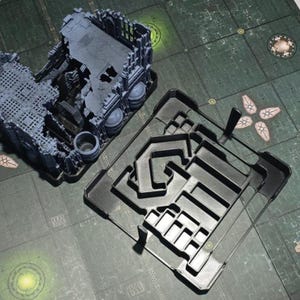 Könnte beinhalten: Ein Tabletop-Gaming-Geländeset mit einem detaillierten, grauen, zerstörten Gebäude und einem schwarzen Kunststoffeinsatz mit Labyrinth-Design. Das Set befindet sich auf einem grün-grauen Spielbrett.