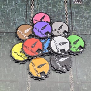 Könnte beinhalten: Eine Sammlung farbenfroher, kreisförmiger Spielmarken mit dem Aufdruck "WOUNDS". Jede Marke hat eine andere Farbe, darunter Rosa, Lila, Grau, Grün, Gelb, Rot, Orange, Braun, Blau und Schwarz. Auf einigen Marken sind Zahlen aufgedruckt.