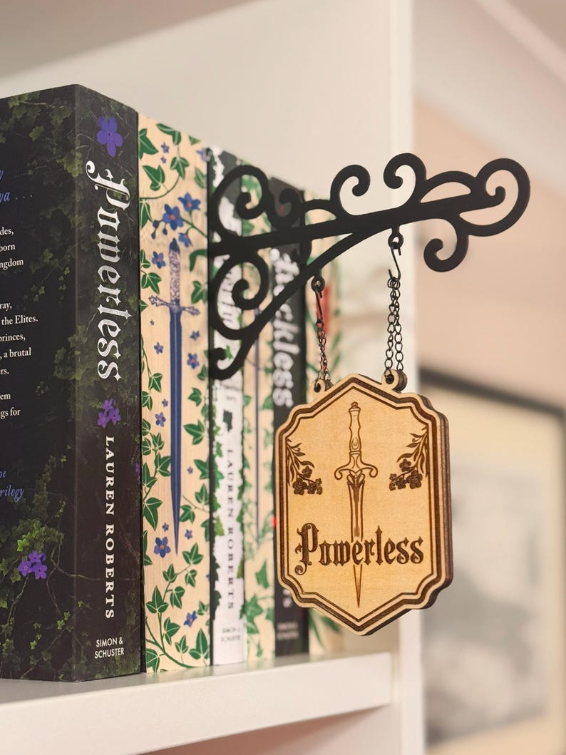 May include: A wooden sign with the word "Powerless" and a sword design hangs from a black metal bracket. The sign is attached to a chain and is in front of a row of books.