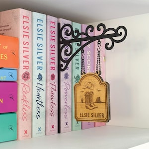 May include: A wooden sign with a cowboy boot and hat design hangs from a black metal bracket. The sign reads "Elsie Silver" and is suspended by a chain. Books with pastel covers are visible in the background.