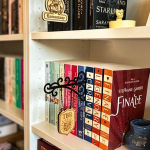 May include: A wooden TBR (To Be Read) sign hangs in front of a row of books on a white bookshelf. The sign is attached to a black metal bracket. The books have colourful spines, and the word "Finale" is visible on one book.