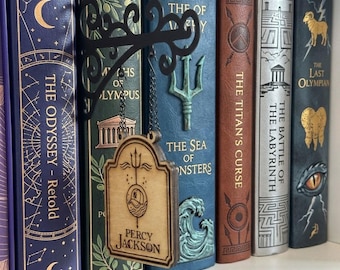 May include: A wooden sign with a laser-engraved trident, a baseball cap, and the text Percy Jackson, suspended by chains from a black metal wall bracket. The sign is positioned in front of a row of hardcover books including The Odyssey and The Last Olympian.