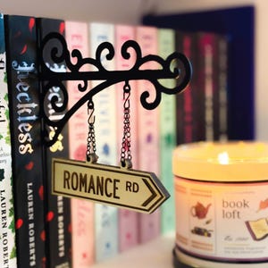 以下が含まれることがあります： 本棚の前に吊るされた「ROMANCE RD」と書かれた黒い金属製の看板。看板は矢印型で、チェーンで吊るされています。背景には「book loft」と書かれた火の灯ったキャンドルがあります。