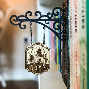 Może przedstawiać: Czarny metalowy uchwyt na znak z dekoracyjnym wzorem przewijania, trzymający drewniany znak z napisem "The Legacy". Znak zawiera grawerowaną laserowo ilustrację postaci. Znak wisi na łańcuchu i jest umieszczony obok rzędu książek.