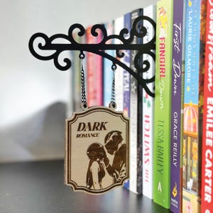 May include: A black metal sign holder with a decorative scroll design, holding a wooden sign that reads "DARK ROMANCE." The sign features an illustration of a couple. Books are visible in the background.