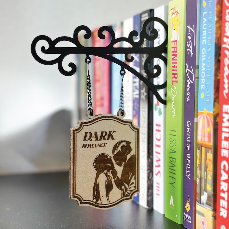 May include: A black metal sign holder with a decorative scroll design, holding a wooden sign that reads "DARK ROMANCE." The sign features an illustration of a couple. Books are visible in the background.