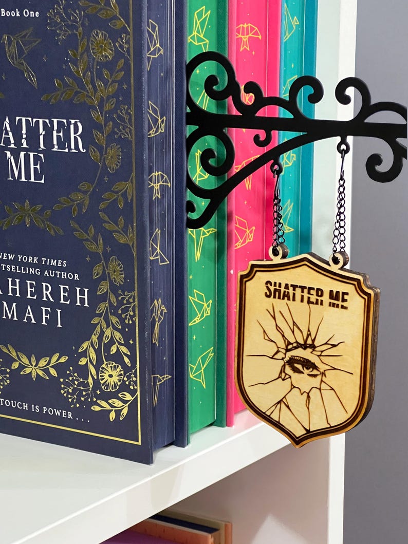 May include: A wooden sign reading "SHATTER ME" hangs in front of a row of colourful books. The sign is shield-shaped with a cracked eye design. A black metal bracket holds the sign, attached to a shelf with books.