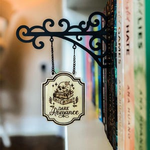 A decorative metal bracket with a hanging sign that reads "Dark Romance" is mounted on a bookshelf filled with various books.