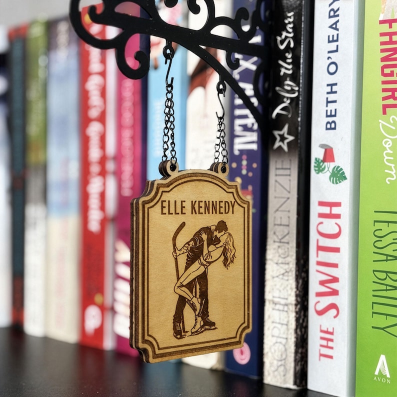 K&ouml;nnte beinhalten: Ein Holzschild mit dem Text "ELLE KENNEDY" und einer Illustration eines Paares beim Eislaufen. Das Schild h&auml;ngt an einer schwarzen Metallhalterung. B&uuml;cher sind im Hintergrund zu sehen.