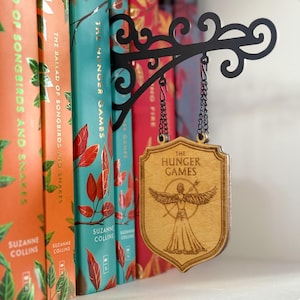 May include: A wooden sign for "The Hunger Games" book series, suspended from a black metal bracket. The sign displays the series title and a graphic of a figure with wings. Colourful book spines are visible in the background.