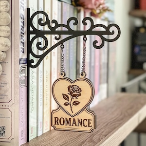 Könnte beinhalten: Ein schwarzes Metall-Buchregal-Schild mit einem herzförmigen Holzschild. Das Schild zeigt eine Rosenillustration und den Schriftzug "ROMANCE". Das Schild hängt über einer Reihe von Büchern.