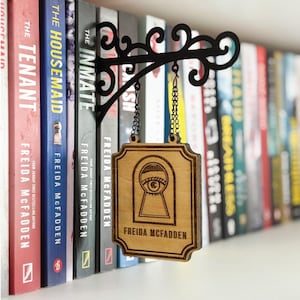 May include: A wooden sign featuring a keyhole design and the text "FREIDA MCFADDEN" is suspended from a black metal bracket. The sign hangs in front of a row of books with colourful spines. The sign is suspended by chains.