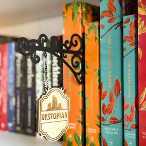 May include: A black metal wall-mounted bracket with a decorative scroll design, featuring a hanging wooden sign suspended by two small chains. The sign is light-coloured wood with a dark engraved illustration of a city skyline and the word DYSTOPIAN.