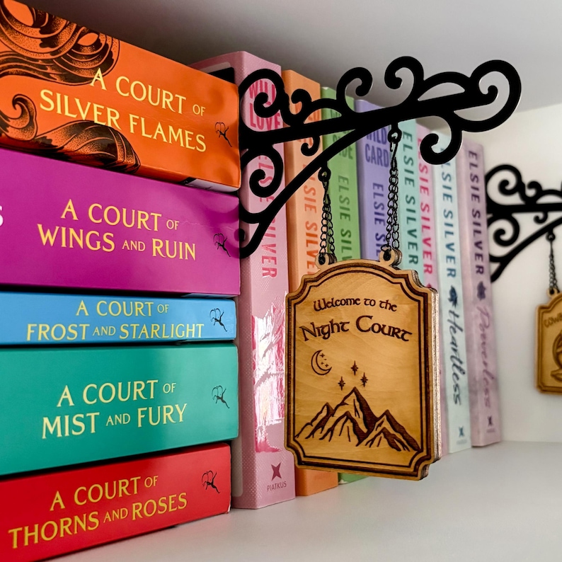 May include: A wooden sign with the text "Welcome to the Night Court" hangs from a black metal bracket. The sign features a mountain range, stars, and a crescent moon design. Books with colorful spines are in the background.