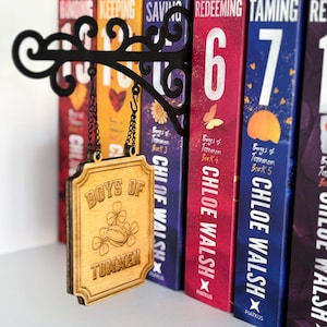 May include: A decorative black metal book holder with a hanging wooden sign that reads "BOYS OF TOMMEN". The sign features a shamrock design. Books with colorful spines are in the background.