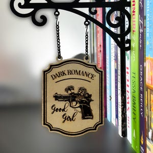 May include: Wooden sign with the words "Dark Romance" and "Good Girl" with a gun and rose design. The sign hangs from a black metal bracket with a chain. A bookshelf with colorful books is in the background.