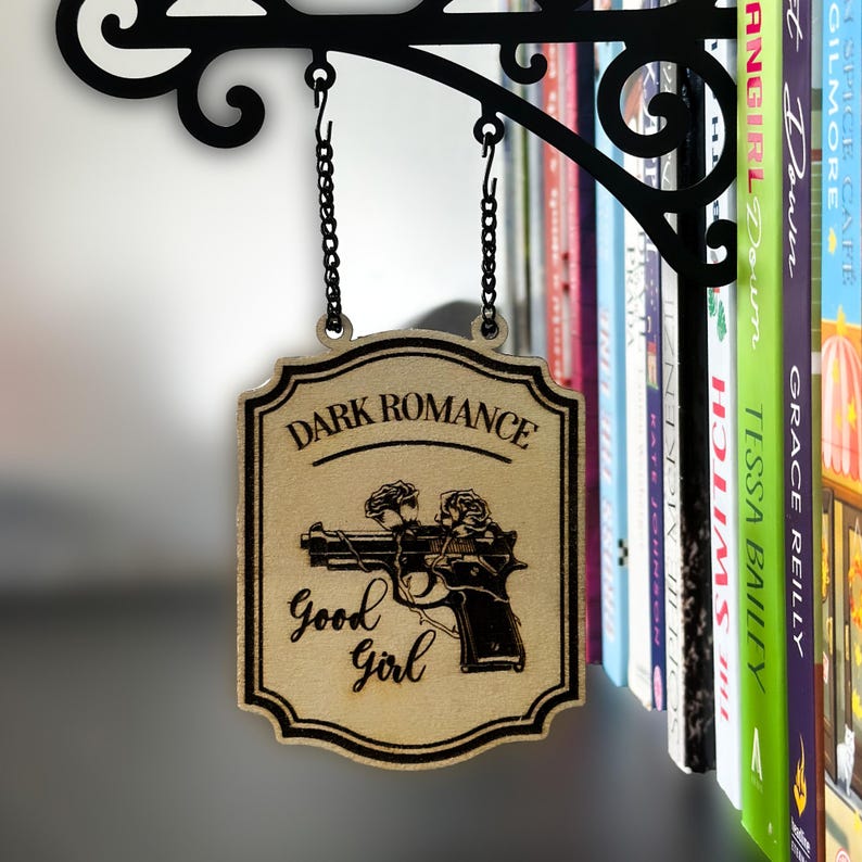 May include: Wooden sign with the words "Dark Romance" and "Good Girl" with a gun and rose design. The sign hangs from a black metal bracket with a chain. A bookshelf with colorful books is in the background.