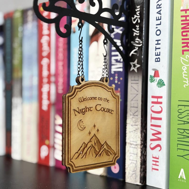 K&ouml;nnte beinhalten: Ein Holzschild mit der Aufschrift "Welcome to the Night Court" h&auml;ngt an Ketten von einer schwarzen Metallhalterung. Das Schild zeigt ein Berg-, Mond- und Sternenmotiv. B&uuml;cher sind im Hintergrund zu sehen.