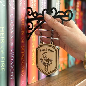 May include: A black metal bookshelf sign with a decorative scroll design is held near a row of books. A wooden shield-shaped pendant hangs from the sign, engraved with the text "Sarah J. Maas" and a deer illustration.