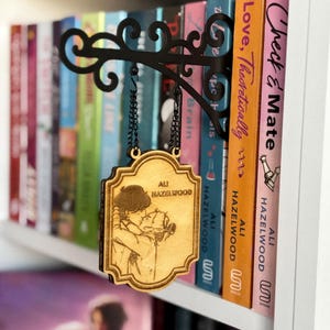 May include: A wooden book sign with a black metal bracket and chain. The sign features an illustration of a couple kissing and the text "ALI HAZELWOOD". The sign hangs in front of a bookshelf with colorful books.