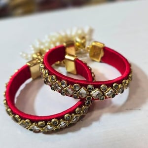 May include: Two red bangles with gold accents and embedded clear gemstones. The bangles are adorned with gold-colored metal and clear, square-cut stones. The jewelry is set against a white background.