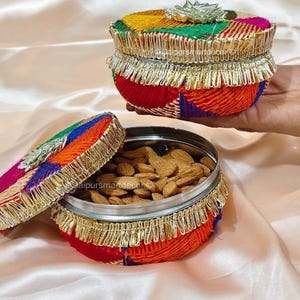 May include: A colorful, round container with a lid, filled with almonds. The container is decorated with vibrant fabric in red, yellow, green, and blue, and trimmed with gold fringe. The lid is open, revealing the contents. Cashews are visible in the foreground.