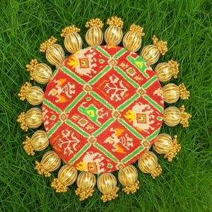 May include: A round textile with a red pattern and green, yellow, and white geometric designs. It is encircled by gold-coloured, beaded ornaments. The image is set against a backdrop of green grass.