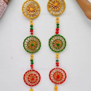 Może przedstawiać: Dwie dekoracyjne dekoracje ścienne z okrągłymi wzorami w kolorze żółtym, zielonym i czerwonym. Każda ma frędzel i akcenty z koralików. Widoczny jest tekst "Jaipursmartdecor".