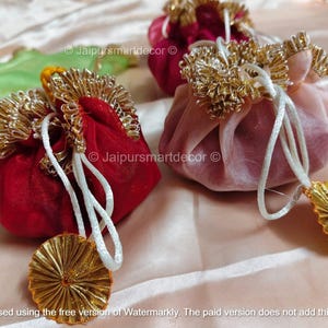 May include: Close-up of several small, round fabric pouches in shades of red and pink, with gold trim and white drawstrings. Each pouch has a gold, fan-shaped tassel. The background is a light pink fabric.