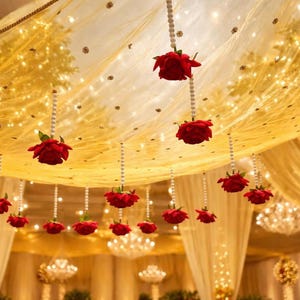 Può includere: Un soffitto decorato con tessuto dorato, luci a catena e rose rosse sospese. Le rose sono attaccate a fili di perline bianche. Lampadari e altre luci illuminano la scena, creando un'atmosfera calda e invitante, perfetta per un matrimonio o un evento speciale.