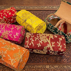 May include: An assortment of colorful, embroidered fabric jewelry boxes and cases. The boxes are in shades of red, yellow, orange, and pink, with gold floral embroidery. One green box is open, revealing red and green bangles.