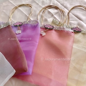 Op de afbeelding: Vier transparante draagtassen in roze, paarse en crèmetinten, met gouden handvatten. Elke tas heeft een decoratieve rand en een lotusbloemaccent. De tassen zijn gemaakt van een licht, doorschijnend materiaal.