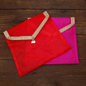 May include: Two fabric envelopes, one red and one magenta. Each has a gold-coloured trim on the flap and a small charm. They are displayed on a dark wooden surface.