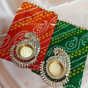 May include: Two decorative candle holders with gold-colored frames and mirrored accents, resting on red and green patterned fabric pouches. The pouches have drawstrings and gold trim.