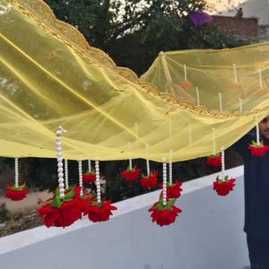 Puede incluir: Una tela amarilla transparente con un borde dorado bordado está adornada con hileras de cuentas blancas y decoraciones de rosas rojas. La tela se sostiene, creando una exhibición decorativa, posiblemente para un evento especial.