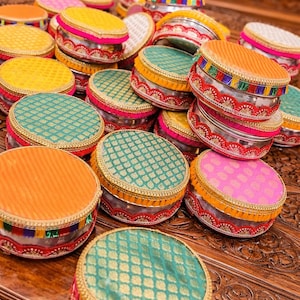 May include: A collection of colorful, round containers with ornate, patterned lids. The lids are in shades of orange, yellow, pink, and teal, with gold trim and decorative edges. The containers are stacked on a wooden surface.