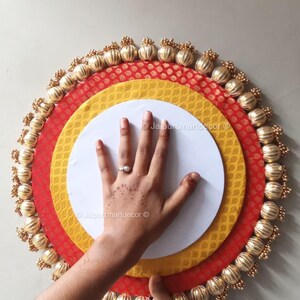 Op de afbeelding: Een decoratief, gelaagd cirkelvormig ontwerp met een wit midden, omringd door gele en rode stoffen met patronen. De buitenrand is versierd met goudkleurige, kralenversieringen. Een hand is gedeeltelijk zichtbaar.