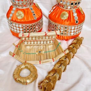 May include: A collection of decorative items. Two orange fabric vases with mirror and gold embellishments, a woven straw basket, a gold ring, and a long, braided tassel are displayed on a light pink fabric.